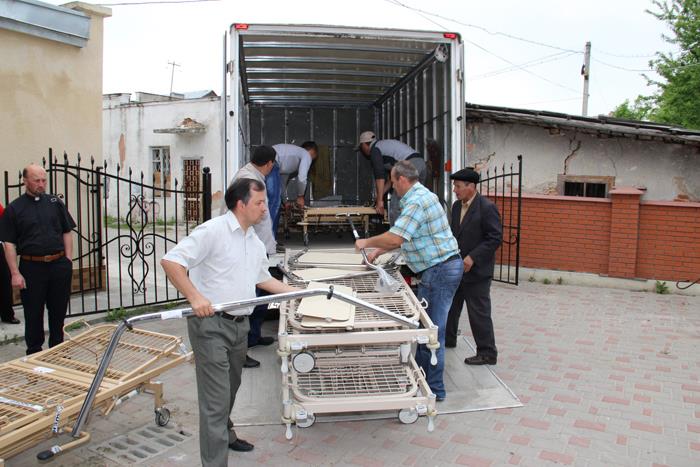 Auf einem großen Platz ist die geöffnete Rückwand eines LKW's zu sehen. Zwei Helfer schieben im Inneren eine Pflegebett nach hinten. Zwei weitere Helfer sind mit einem schon abgeladenen  Pflegebetten 