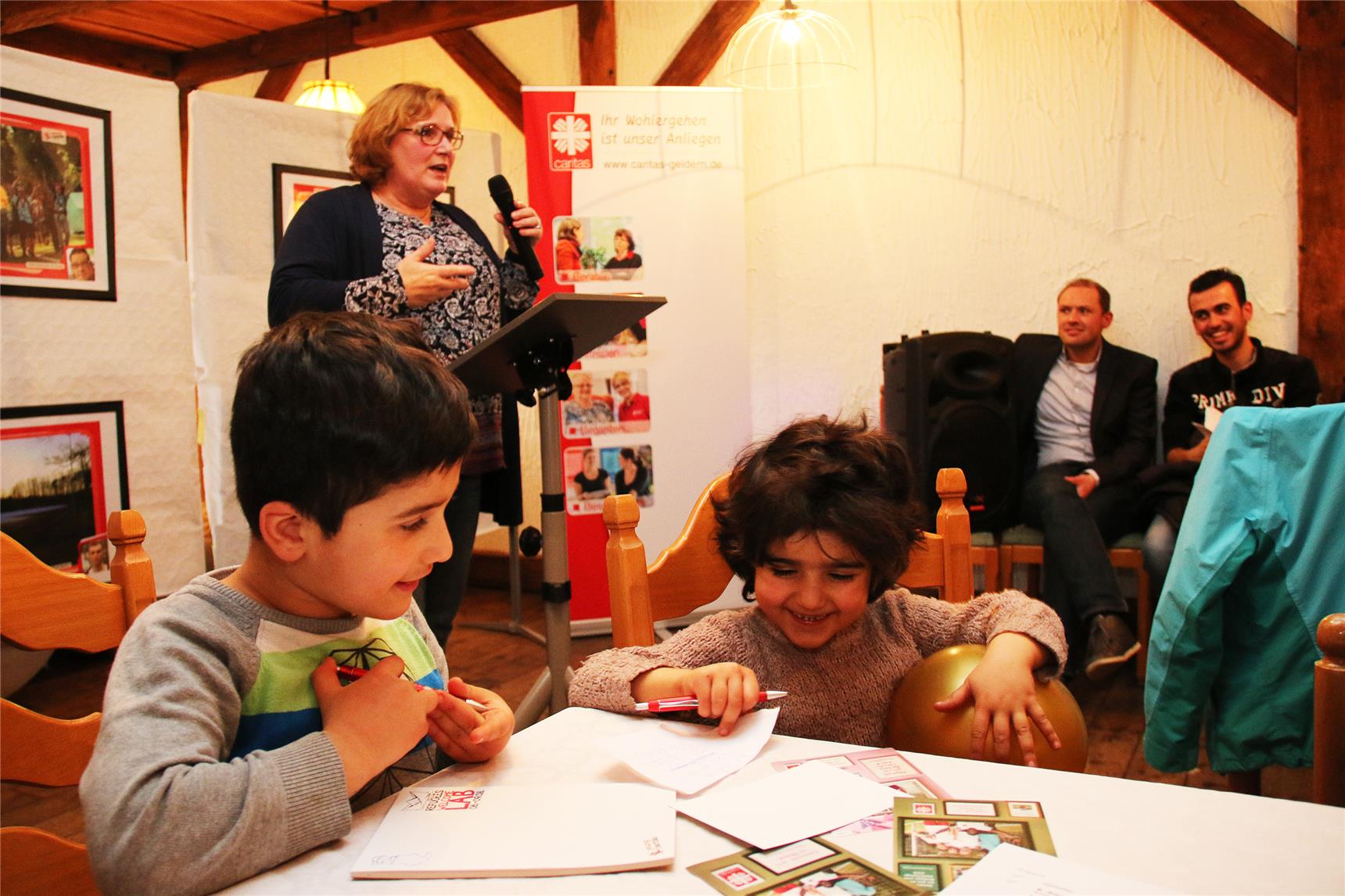 Zwei Kinder sitzen am einem Tisch und malen und eine Frau redet im Hintergrund.