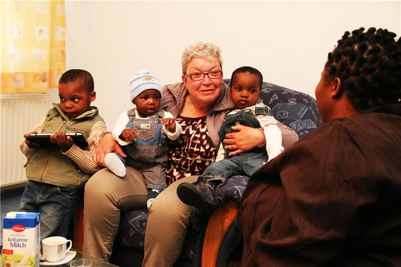 Eine Frau sitzt auf einem Sofa und hat zwei Kleinkinder auf dem Schoß, eins steht daneben