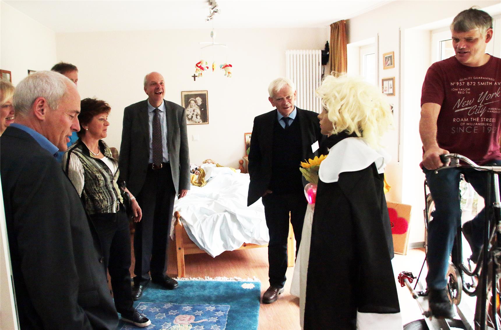 Das Foto zeigt einen Mann auf einem Fahrrad in einem Zimmer und eine kleine Gruppe von Menschen, die ihm zuschauen.