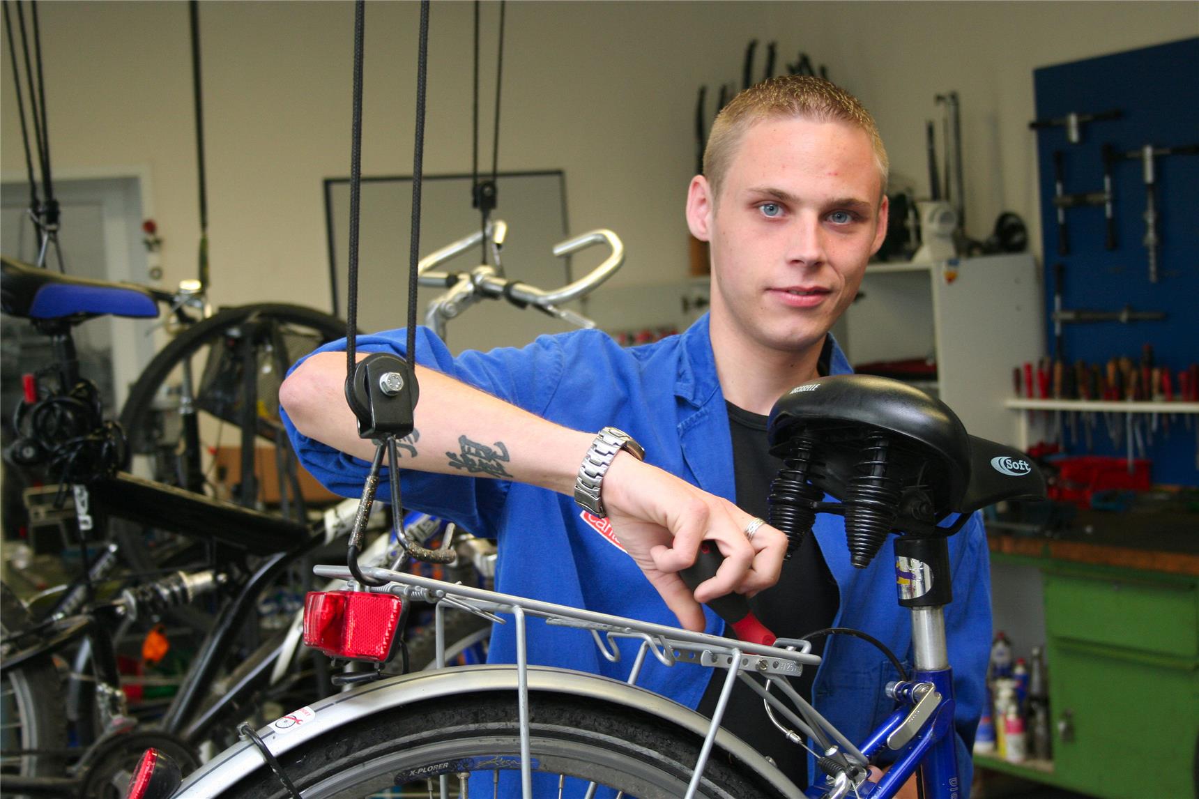 Das Foto zeigt einen jungen Erwachsenen in blauer Arbeitskleidung, der an einem aufgehängten Fahrrad schraubt.