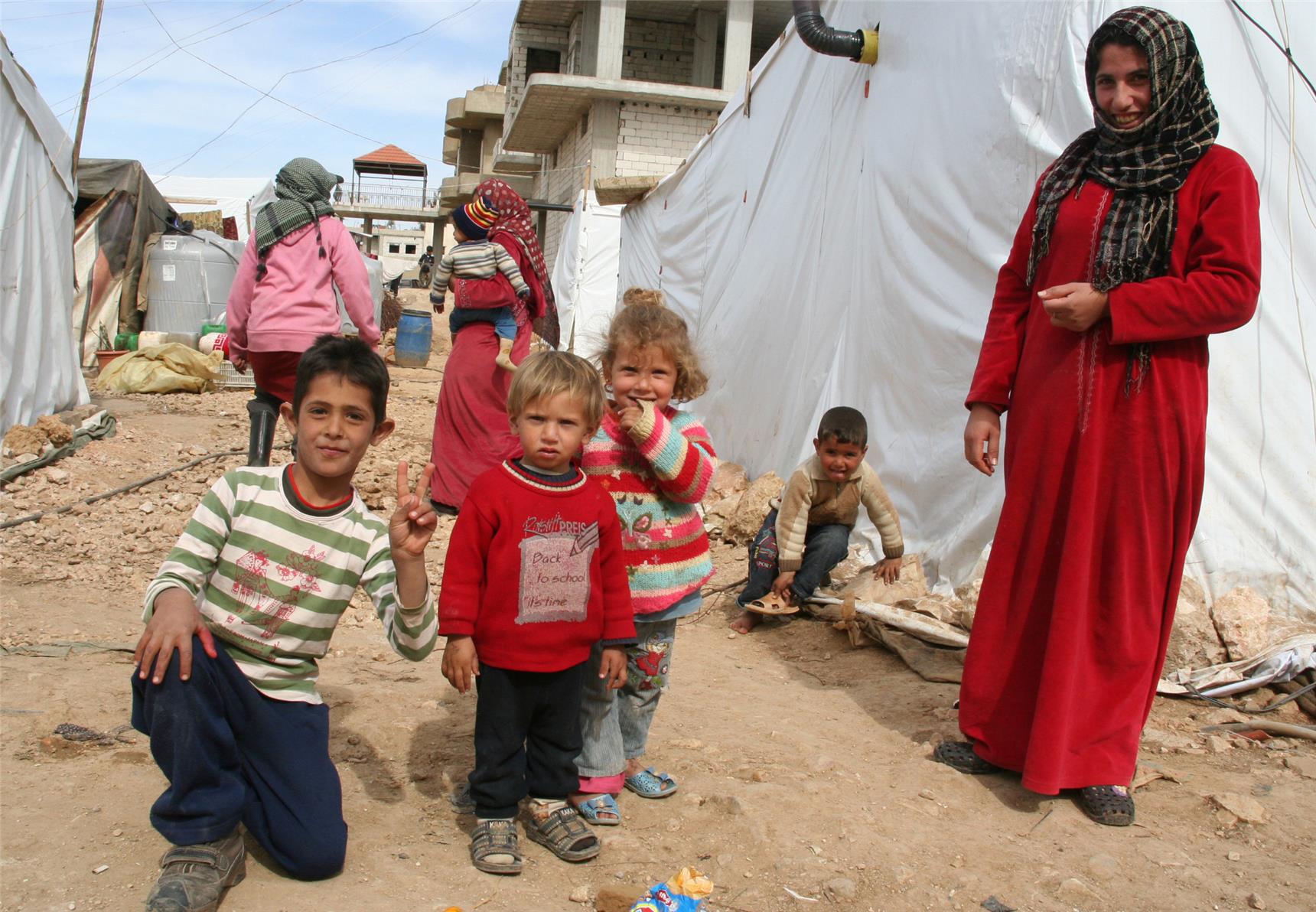 Drei kleine Kinder stehen auf trockenem Boden und schauen in die Kamera. Das Flüchtlingslager besteht aus notdürftig errichteten Unterkünften. Zelthütten aus weißer Plane sind mit Steinen befestigt.