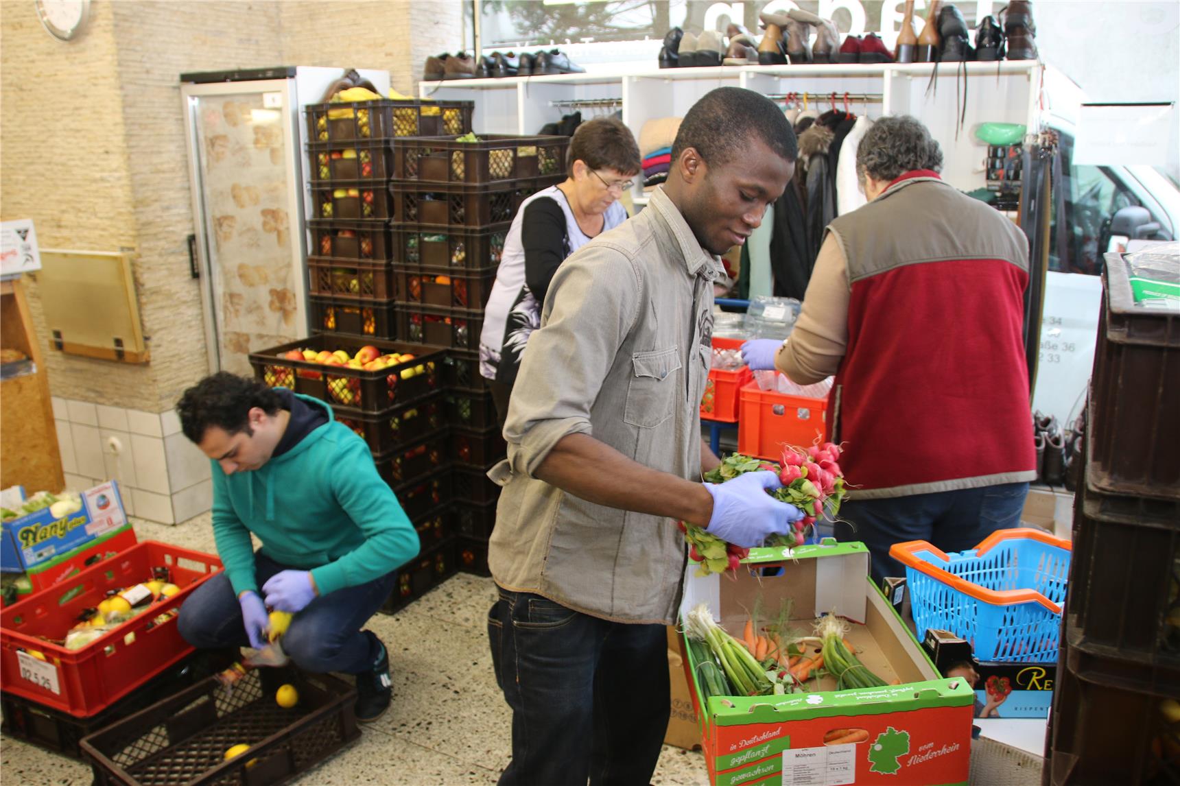 Das Foto zeigt einen jungen Flüchtling, der Gemüse in einer Kiste sortiiert, im Hintergrund weitere Mitarbeitende der Tafel, die den Verkauf vorbereiten.