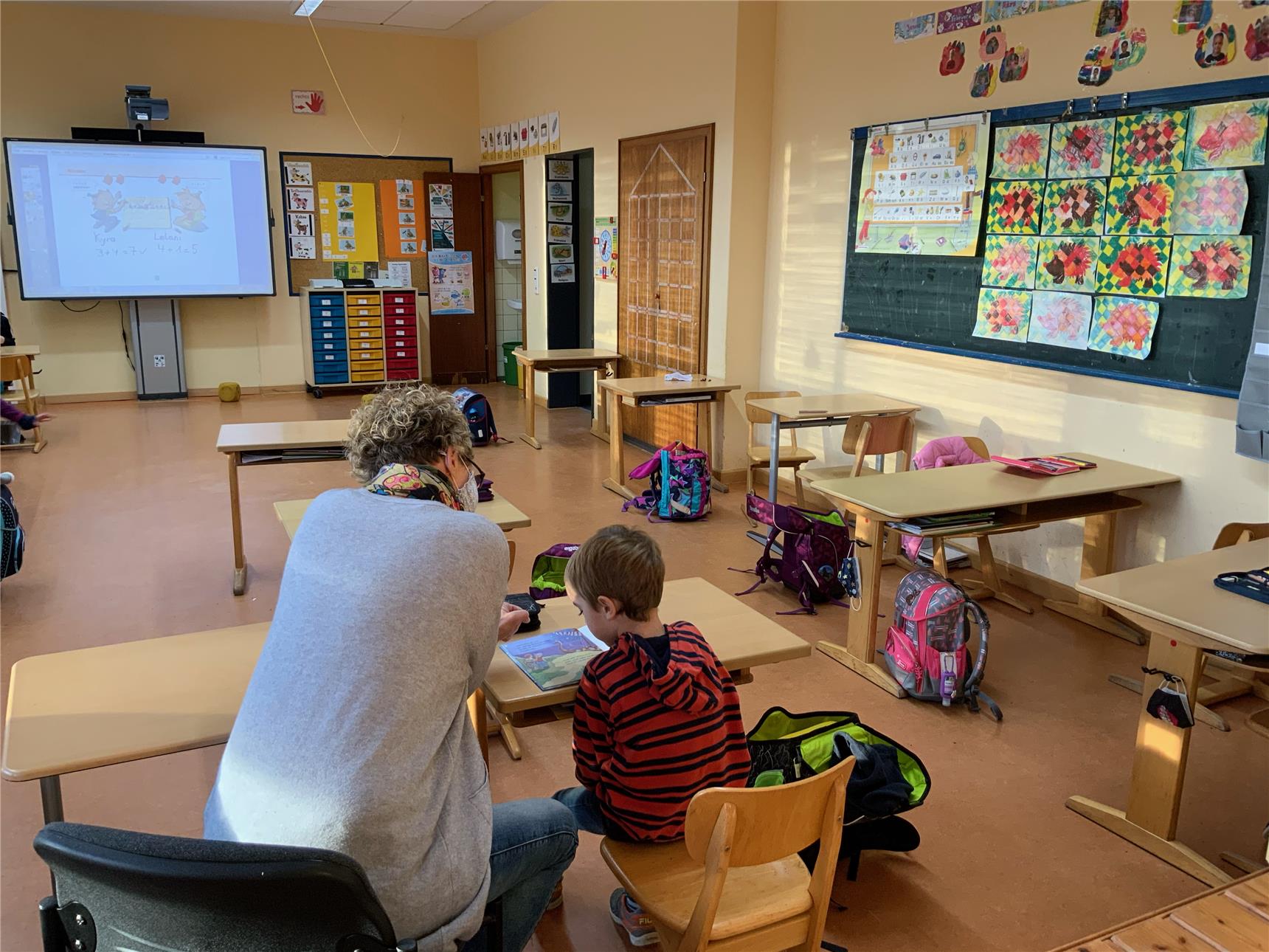 Das Foto zeigt eine Frau, die mit einem Jungen in einem Klassenraum sitzt, von hinten.