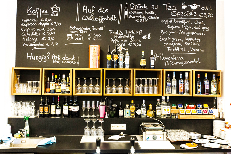 Die Theke der Bar des Hotel 'Magdas' in der Frontansicht. Es sind die Theke, Regale mit Gläsern und Getränken sowie eine Tafel mit der Getränke- und Speisekarte zu sehen.