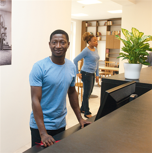 Ein junger Mitarbeiter der Empfangstresen des Wiener Hotels 'Magdas' steht und in die Kamera blickt. Im Hintergrund steht eine weitere Mitarbeiterin.