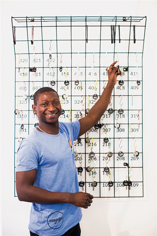 Ein Mitarbeiter des Hotel 'Magdas' steht vor einem Schlüsselbrett, greift nach einem Zimmerschlüssel und blickt dabei lächelnd in die Kamera