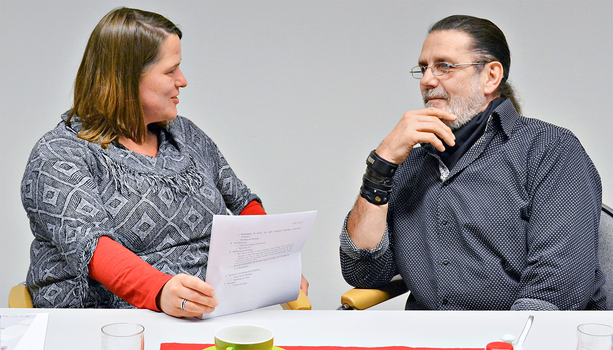 Ein weiblicher Coach und ein Lotse des Projektes 'Schritt für Schritt' sitzen an einem Tisch reden miteinander. Der Coach hat Dokumente in der Hand.