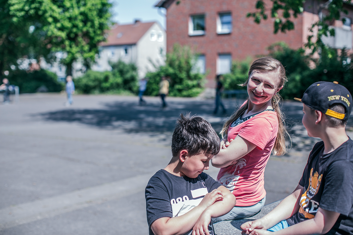 Die Betreuerin einer OGS lehnt bei Sonnenschein an einer Tischtennisplatte auf einem Schulhof. Neben ihr befinden sich zwei Jungen. Im Hintergrund sind weitere Kinder auf dem Schulhof zu sehen.