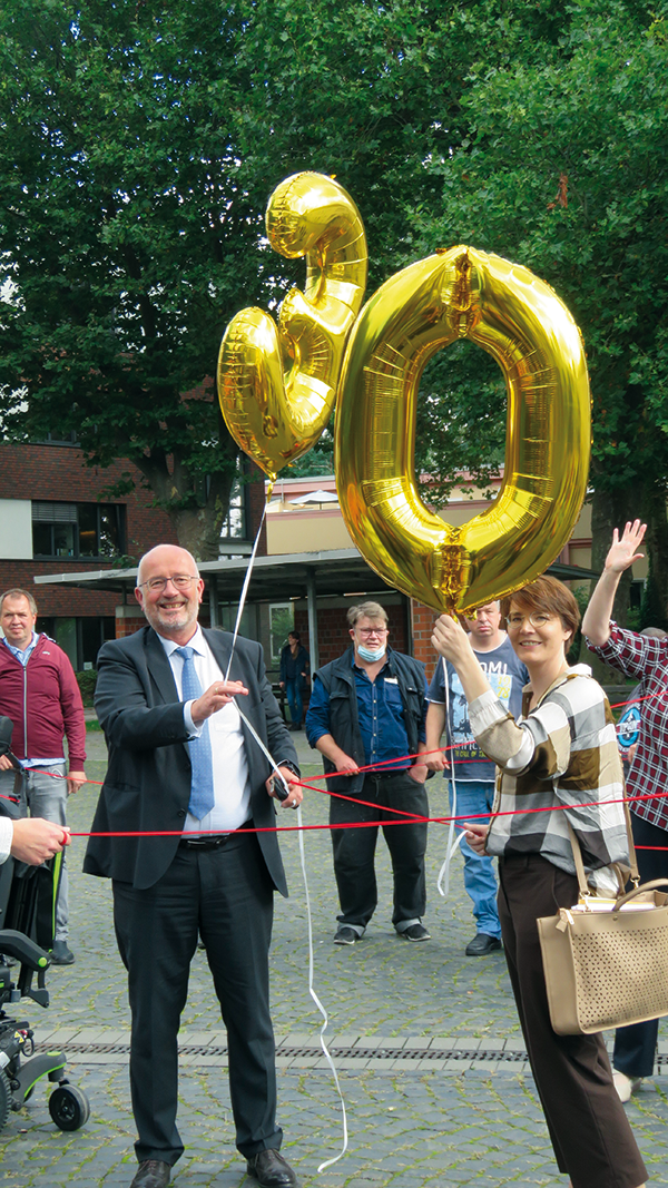 Der Paderborner Diözesan-Caritasdirektor Josef Lüttig hält zusammen mit einer Frau zwei goldenen Luftballon, die die Zahl 30 darstellen, in der Hand. Zwischen ihnen wurde eine rote Schnur gespannt.