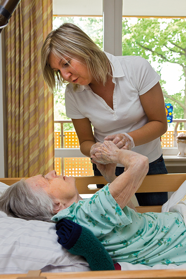 Eine Pflegerin steht an einem Krankenbett, in dem eine Seniorin liegt. Beide halten sich an den Händen und blicken sich in die Augen.