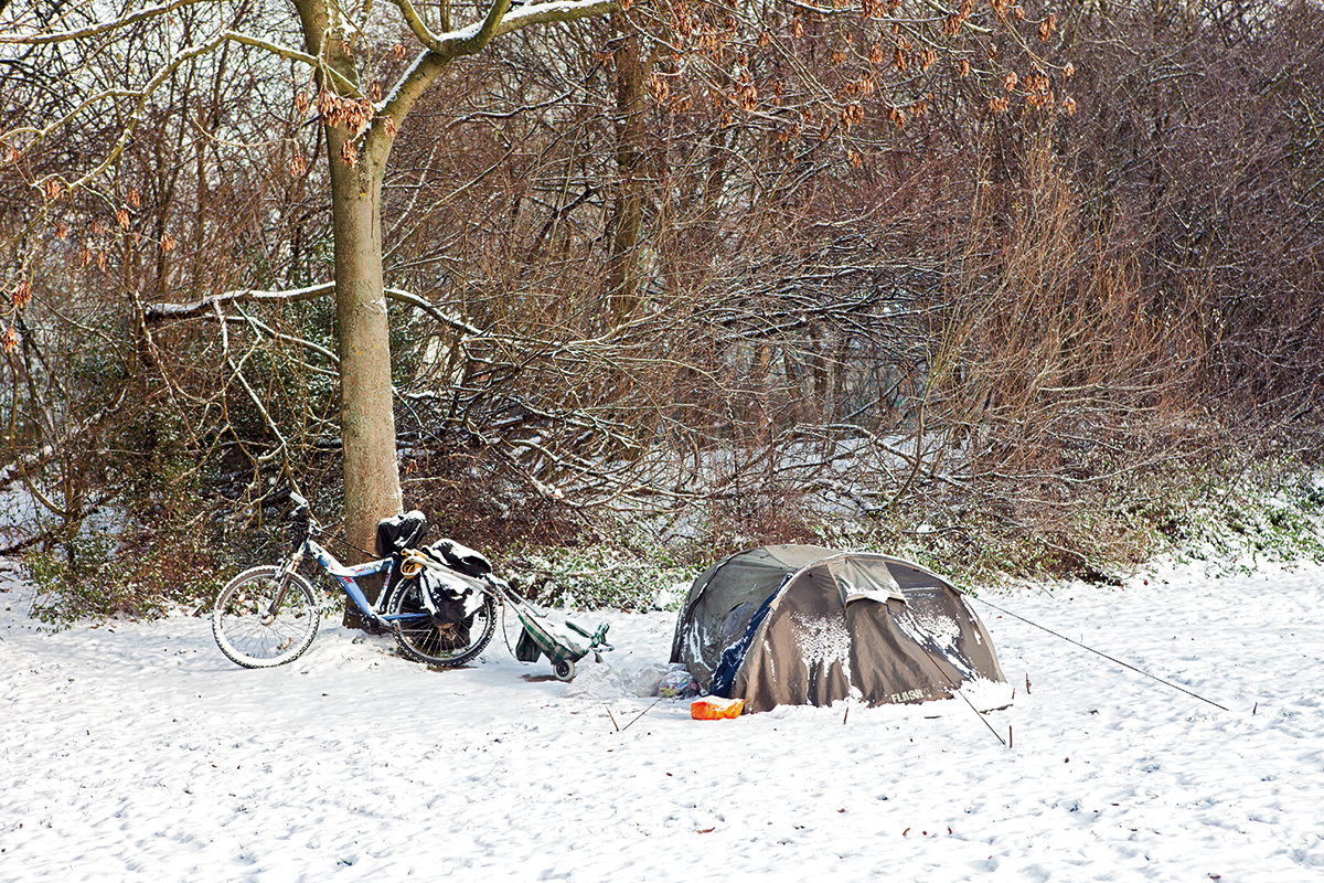 Der Schlafplatz eines Obdachlosen mit aufgebautem Zelt, Fahrrad und Einkaufkarre, der sich auf einer mit Schnee bedeckten Wiese vor einem Waldstück befindet
