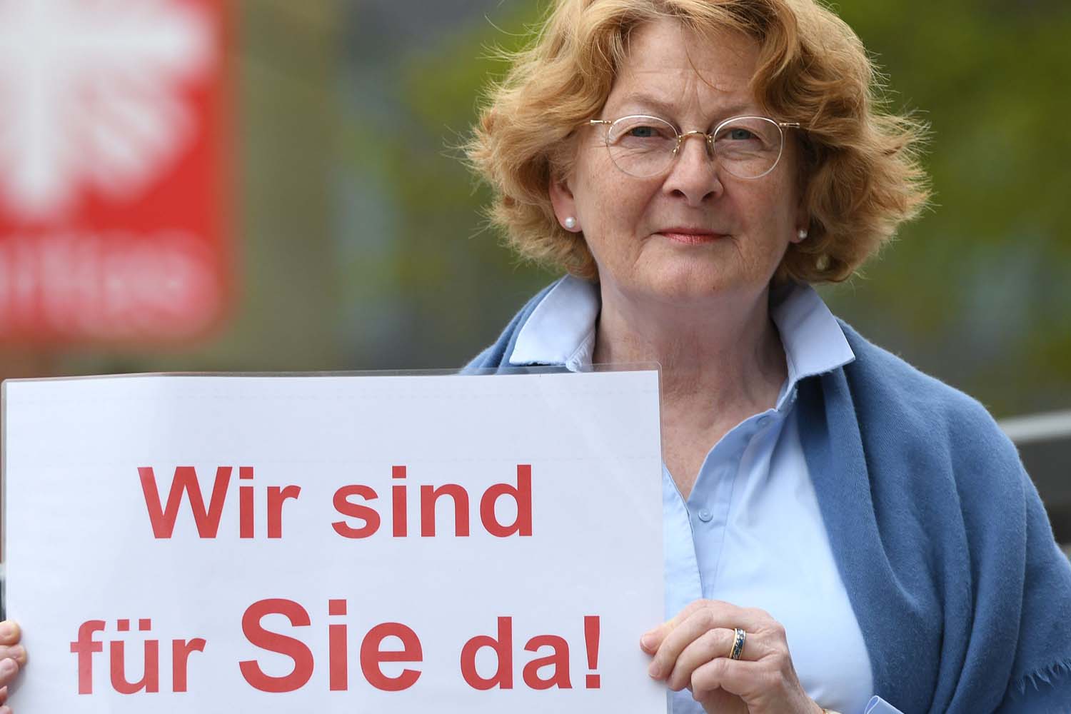 Eine ältere Dame mit Brille hält ein Schild hoch auf dem steht: Wir sind für Sie da.