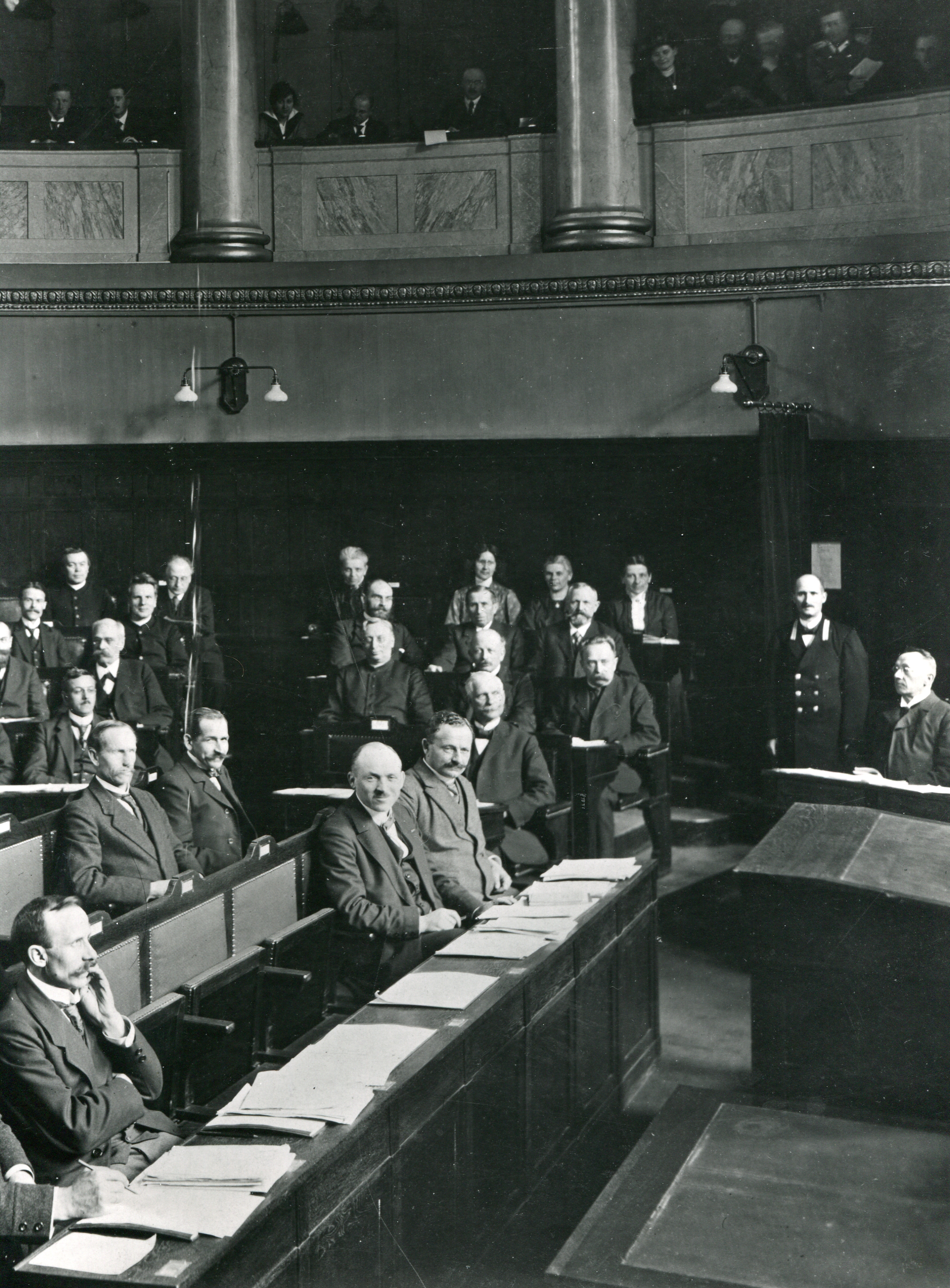 Schwarz gekleidete Menschen sitzen in einem Saal, einige davon stehen