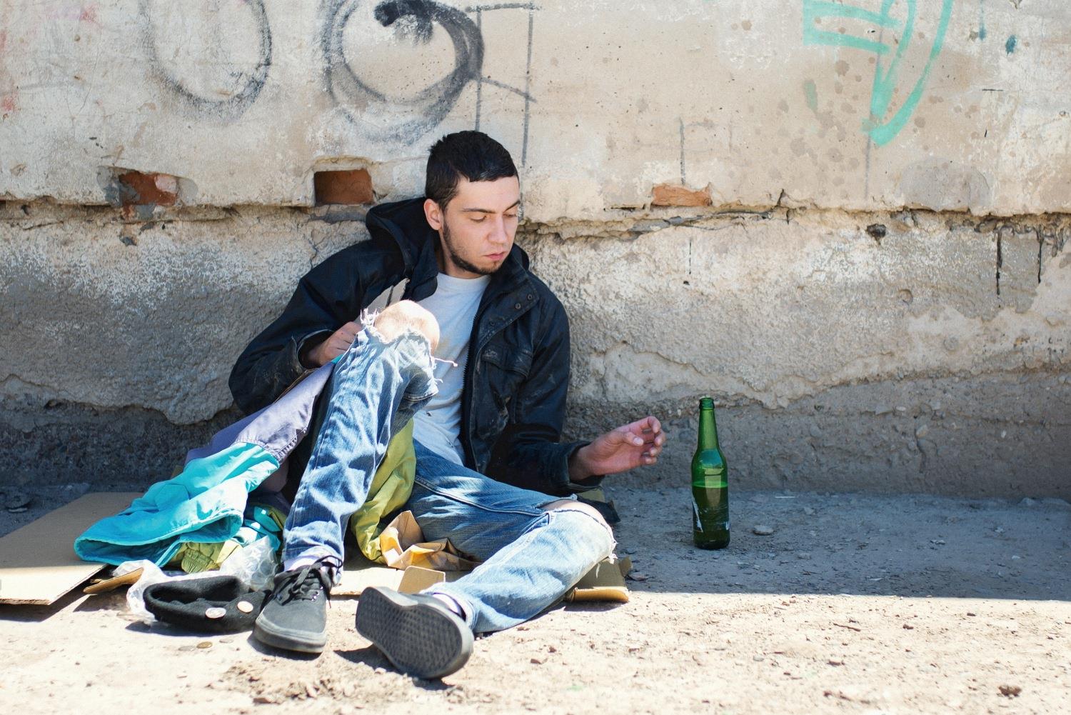 Junger Mann, der auf der Straße auf Pappkarton sitzt, greift nach einer Bierflasche.