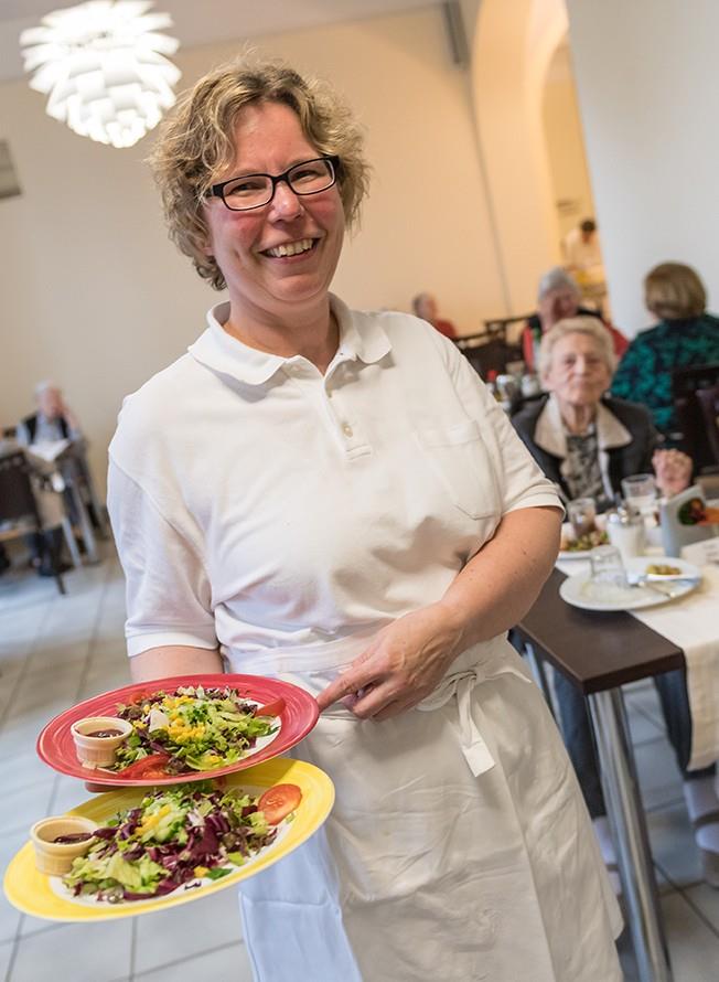 Eine sympathische Hauswirtschafterin bringt Essen in den Speiseraum eines Altenheims. Im Hintergrund Bewohnerinnen an ihren Tischen.