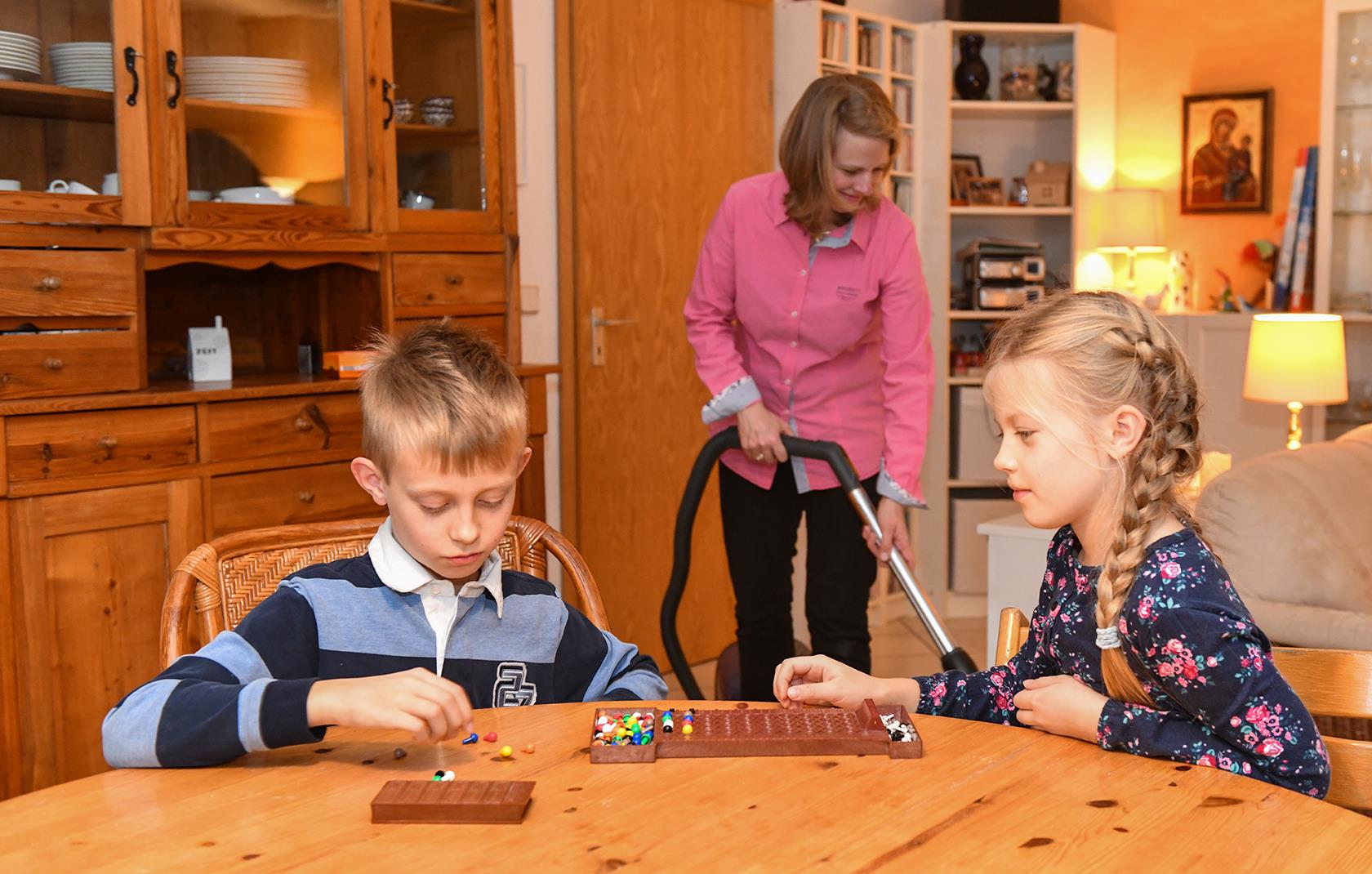 Zwei Kinder sitzen am Tisch und im Hintergrund saugt eine Frau Staub