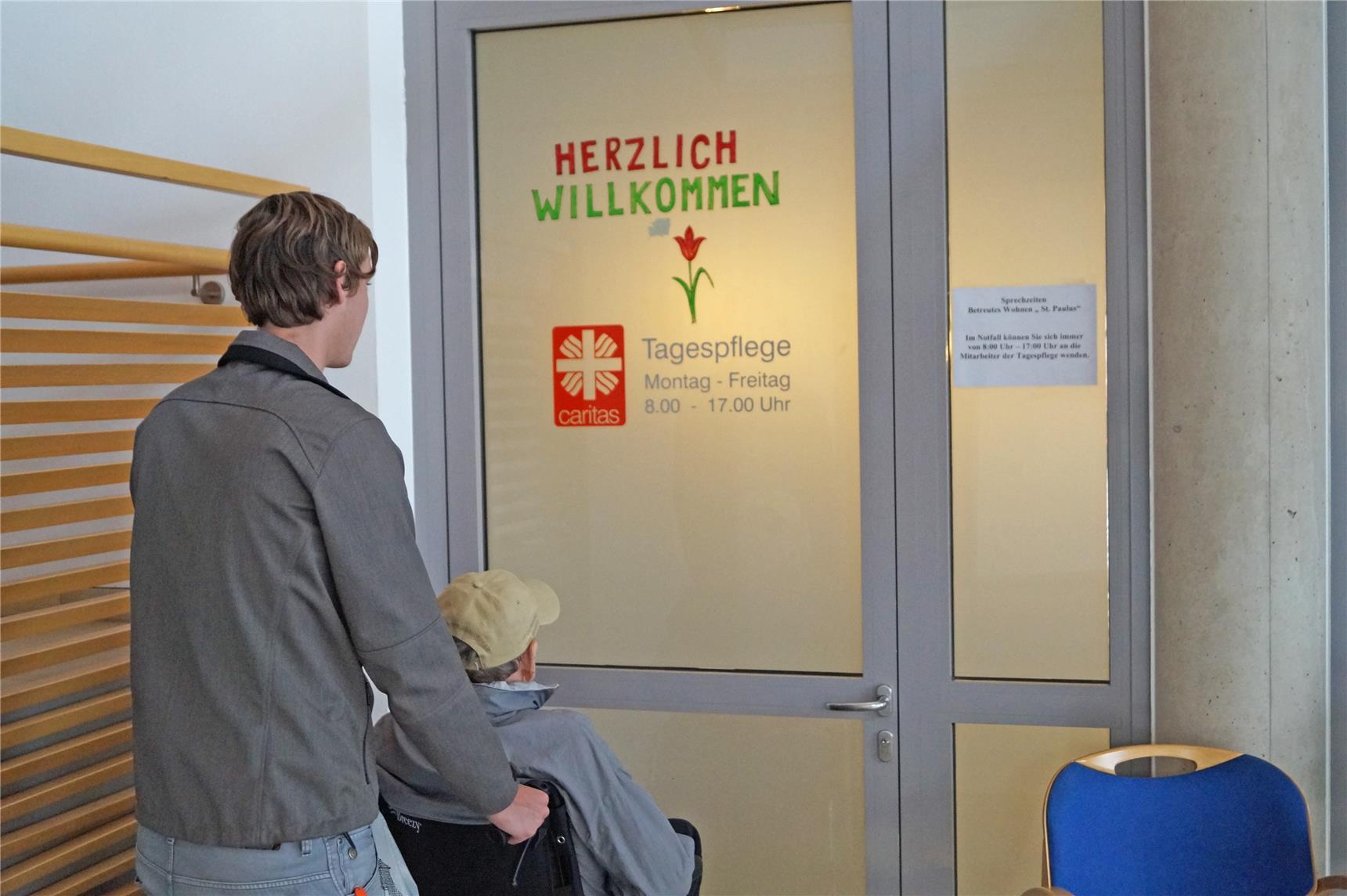 Person im Rollstuhl und Pfleger vor Eingang zur Tagespflege