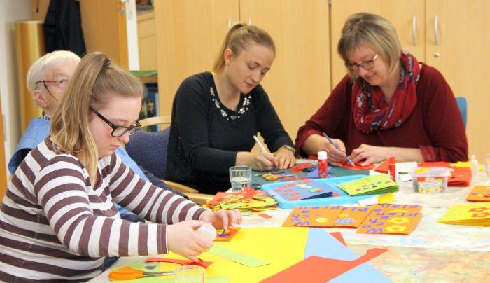 Senioren, Kinder und Betreuer sitzen an einem Tisch und basteln mit Papier.