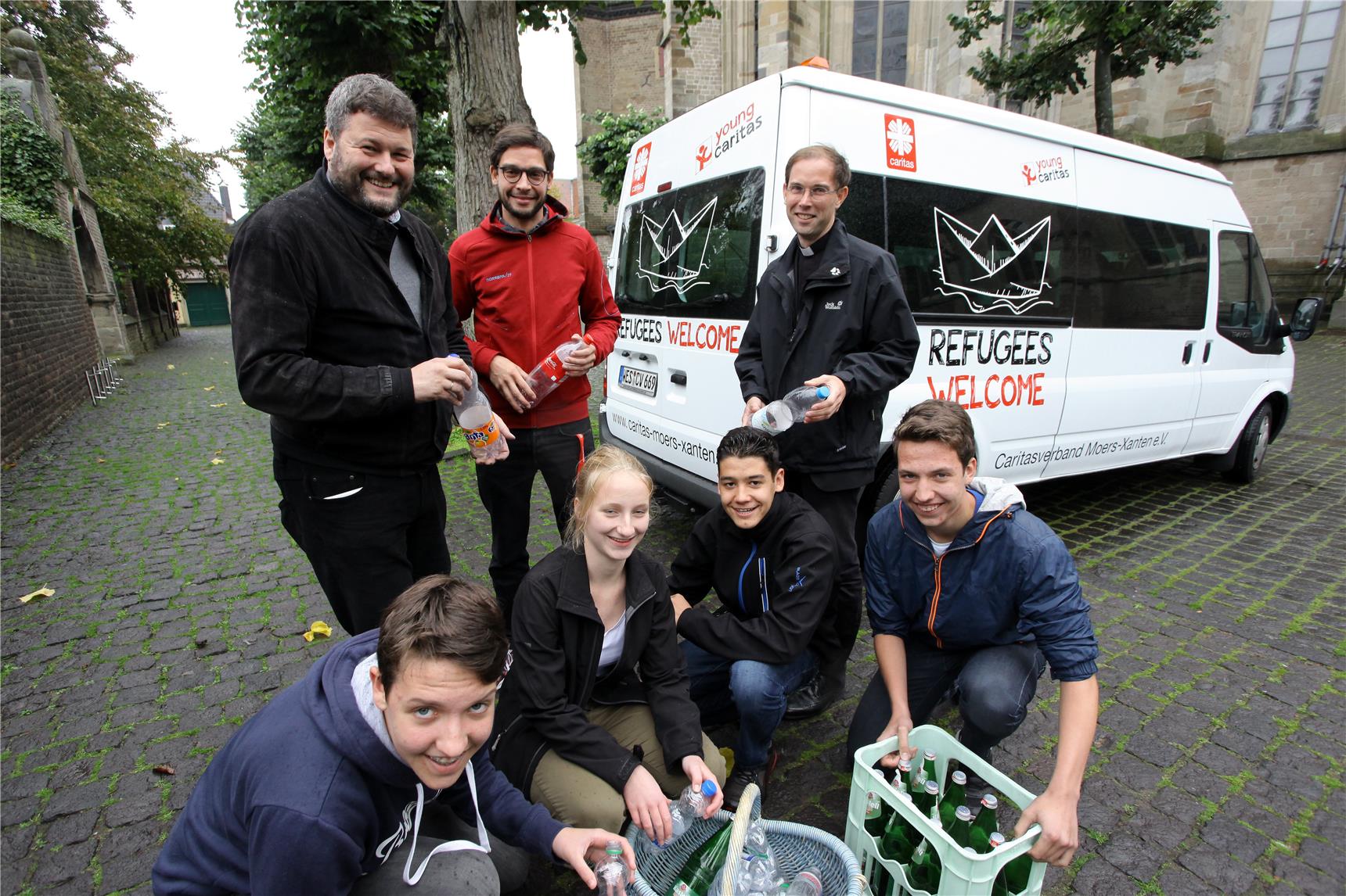 Auf dem Bild stehen und hocken einige Jugendliche mit Pfandflaschen vor einem Caritas-Bulli