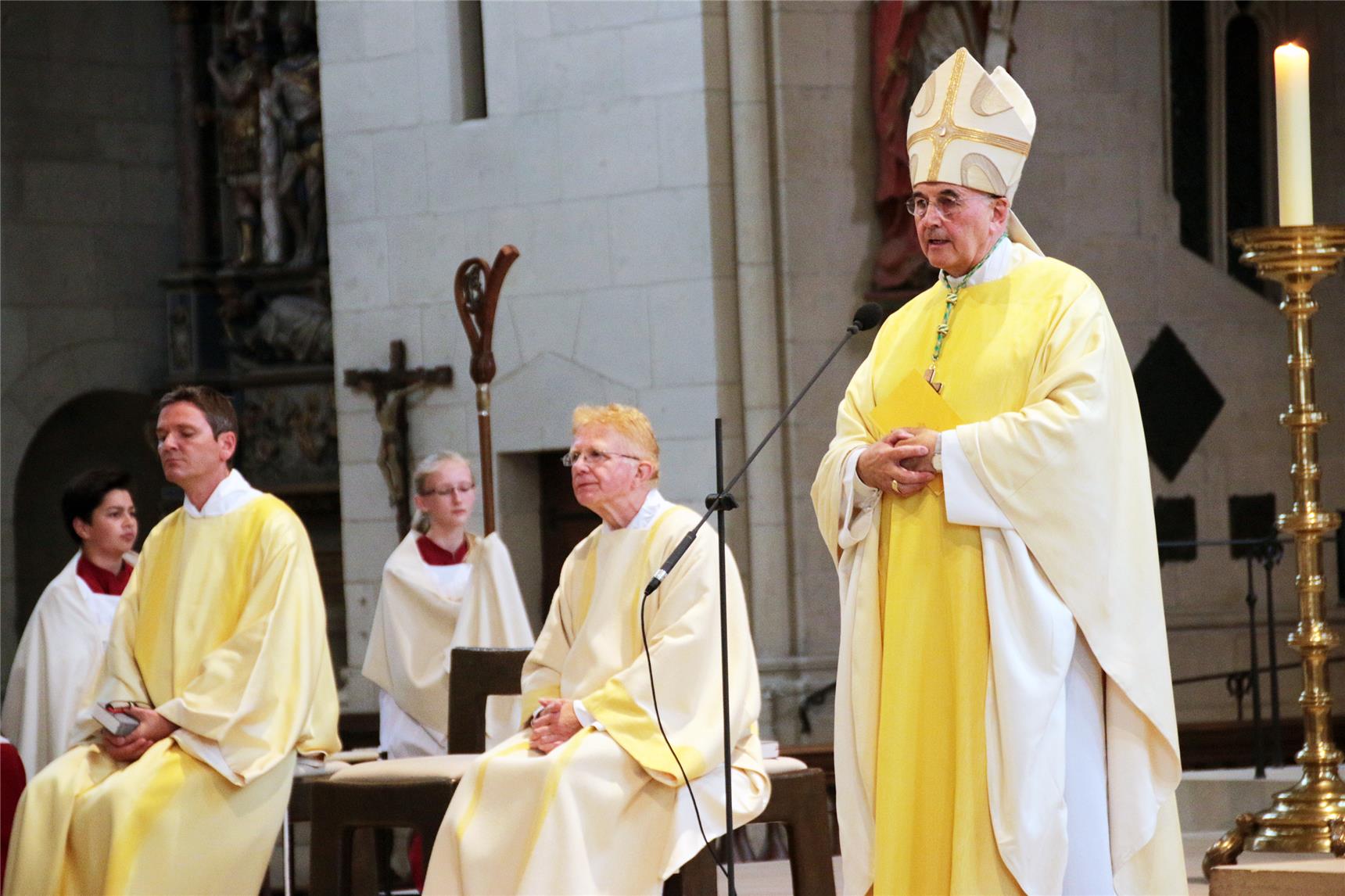 Auf dem Bild sitzen mehrere Geistliche, der Bischof steht am Mikrofon