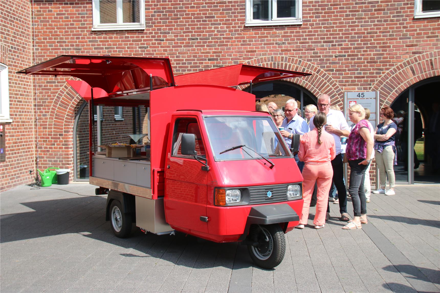 Auf dem Bild stehen mehrere Menschen vor der roten Ape