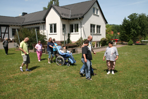 Hier ist die Gruppe vor einem weißen Haus mit grauem Dach auf der Wiese. Alle sind in Bewegung.  Sie