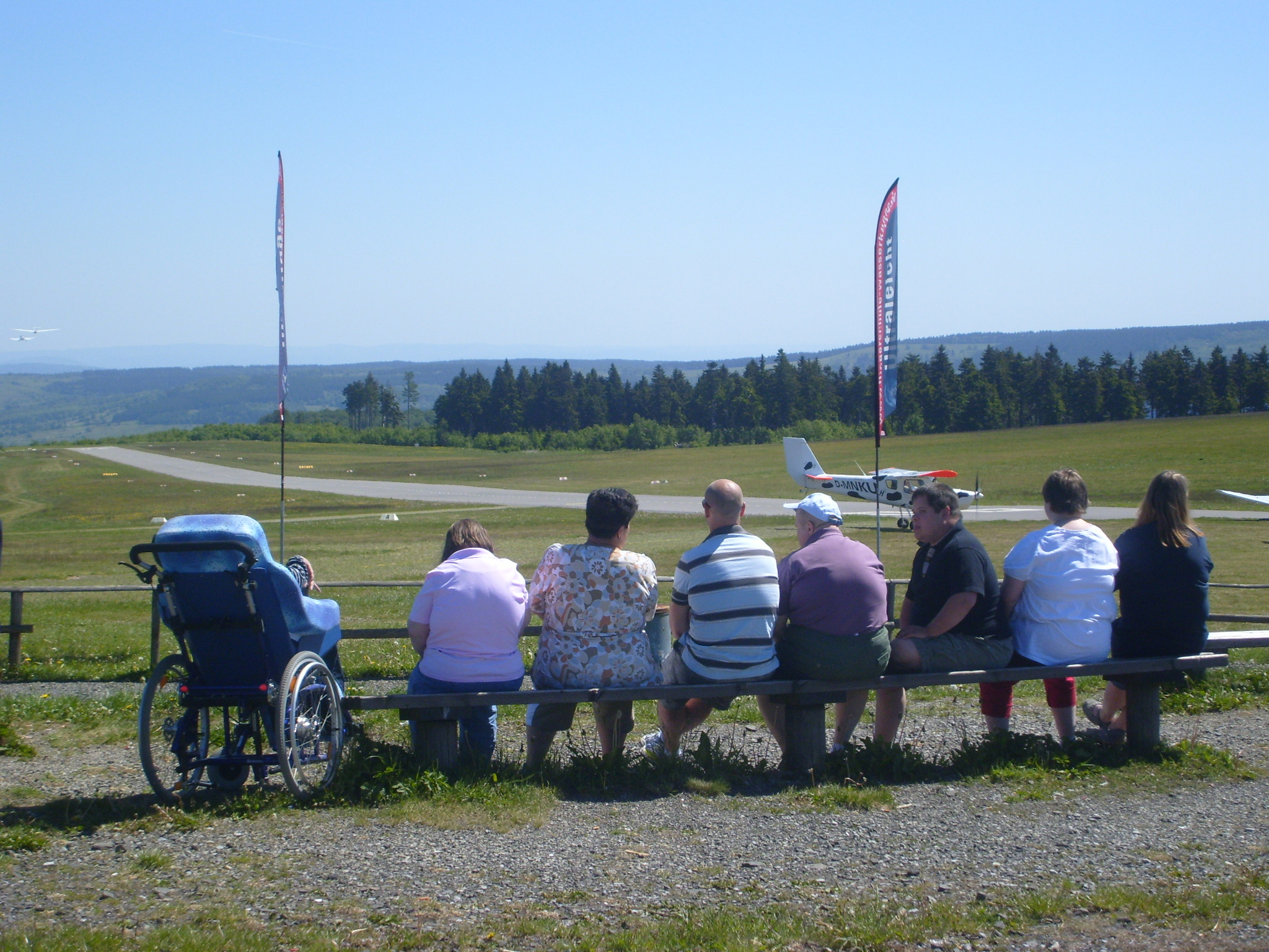 Die Gruppe sitzt auf einer Bank vor dem Flugplatz auf der Wasserkuppe. Alle schauen gespannt dem Flu