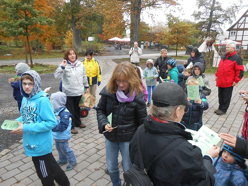 Auf einem gepflasterten Platz am Waldrand haben sich Kinder und Erwachsene eingefunden. Viele von ihnen halten eine grüne Proschüre in ihren Händen. Einige haben sie aufgeschlagen und lesen darin. Ein
