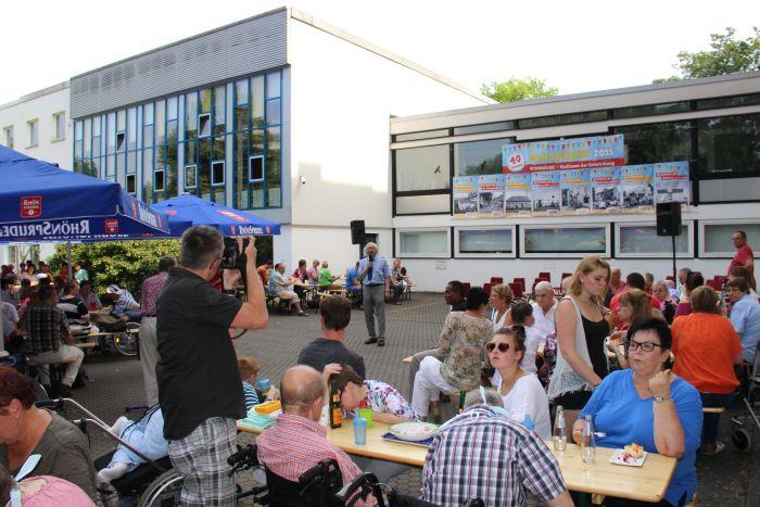 In der Mitte des Festplatzes steht Herr Chrome und spricht zu dem Anwesenden. Im Hintergrund sehen wir das gelb verkleidete Gebäude der Behindertenhilfe in der Ratgarstraße in Fulda.