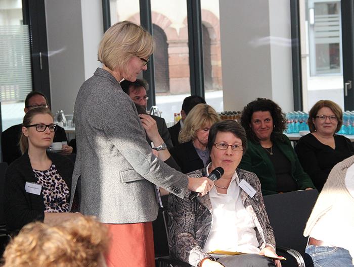 Zwischen den Teilnehmern steht eine Frau mit einem Mikro und hält es einer Teilnehmerin zum Sprechen bereit.