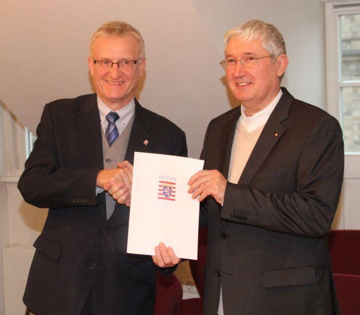 Beide Herren geben sich die rechte Hand und halten den Bescheid vor sich. 