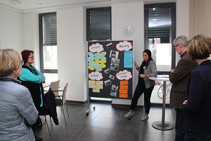 Eine Frau steht vor einer Pinwand und spricht zu dem Teilnehmern. 