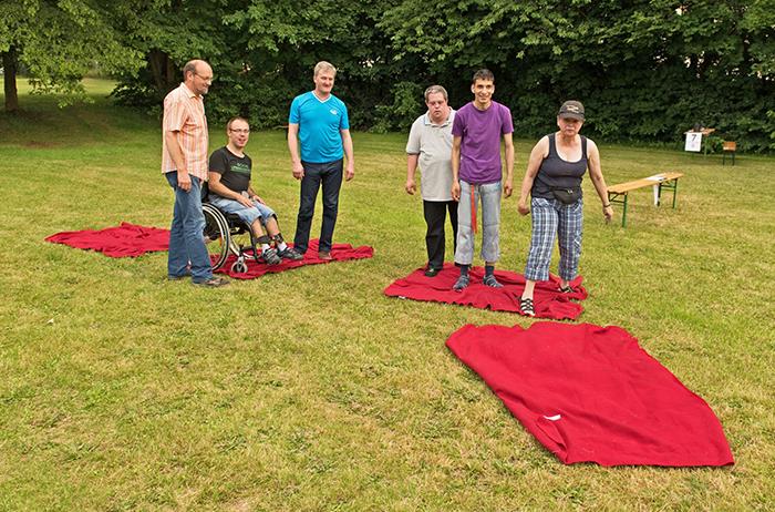 Auf der grünen Wiese sind rote Decken als begehbare Flächen ausgelegt. Jedes Team muss sich von einer Decke zur anderen fortbewegen.