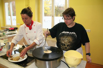 Eine Frau läst Teig aus einer Schöpfkelle auf eine heiße Kochplatte fließen. 